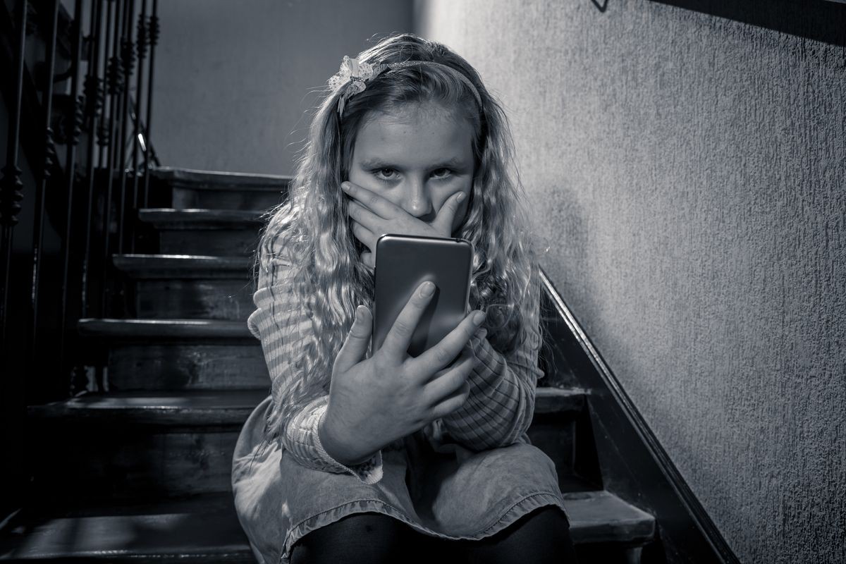 Sad depressed young girl victim of cyberbullying by mobile smart phone sitting on stairs feeling lonely, unhappy, hopeless and abused. Child bullied and harassed by text message by online stalker.